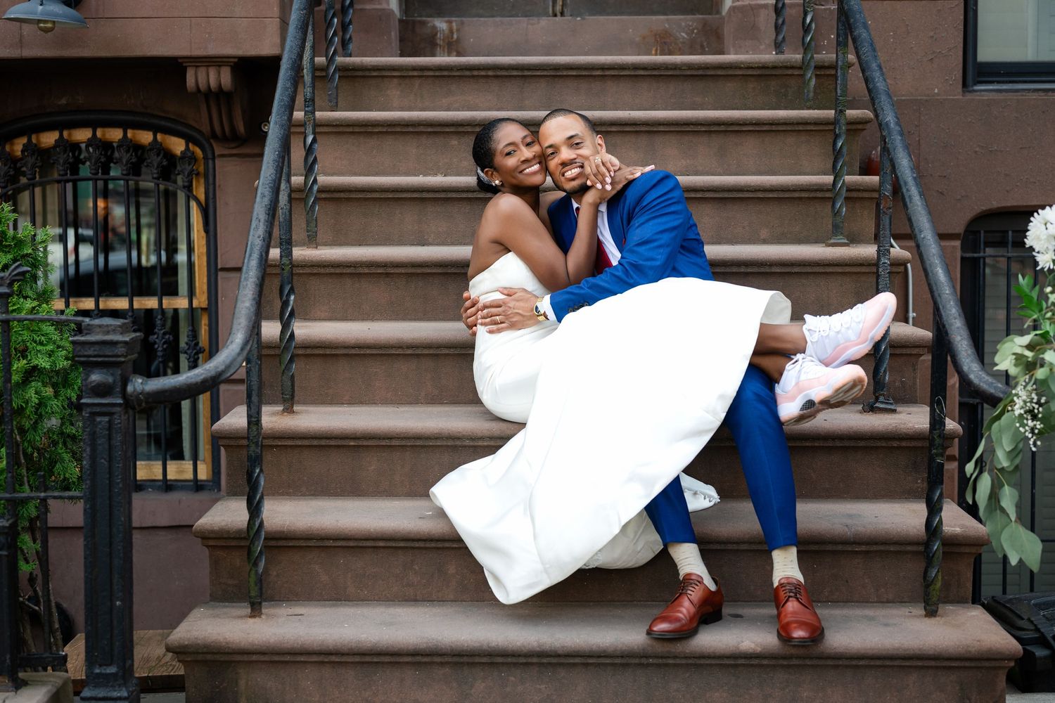 A couple in wedding attire share a romantic moment on brownstone steps outside a urban building.