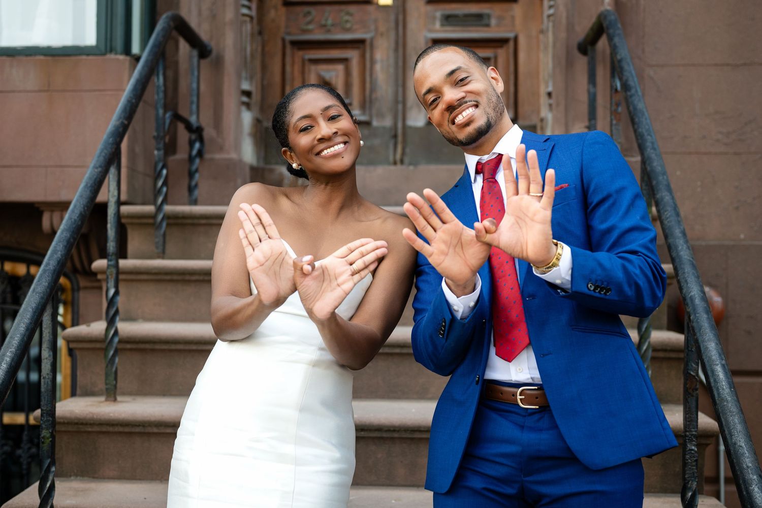 A newlywed couple shows off their wedding rings while standing on brownstone steps, with bride in white and groom in blue suit.