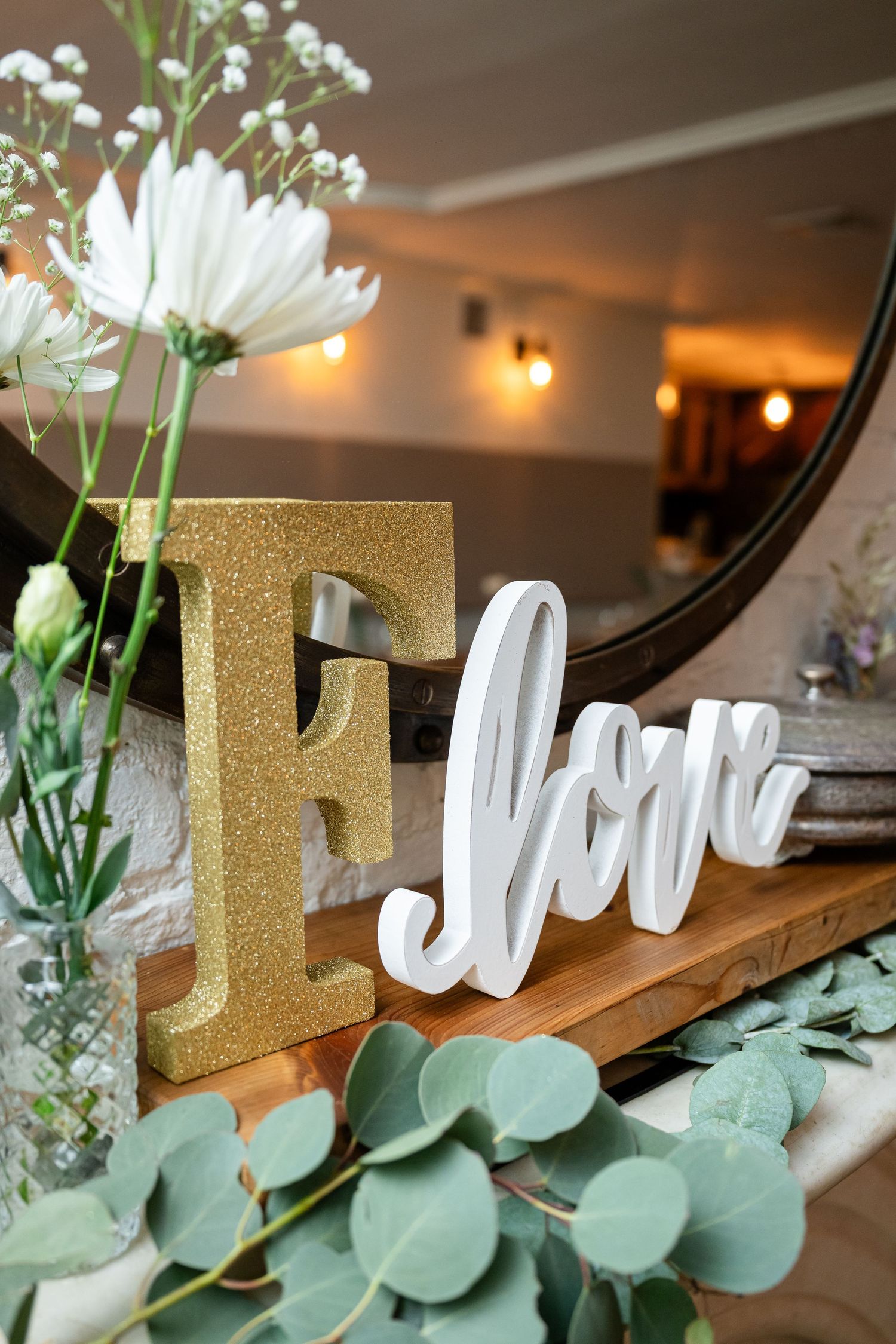 Wedding decor featuring a gold letter F and white 'love' sign with fresh eucalyptus and white flowers on a wooden shelf.