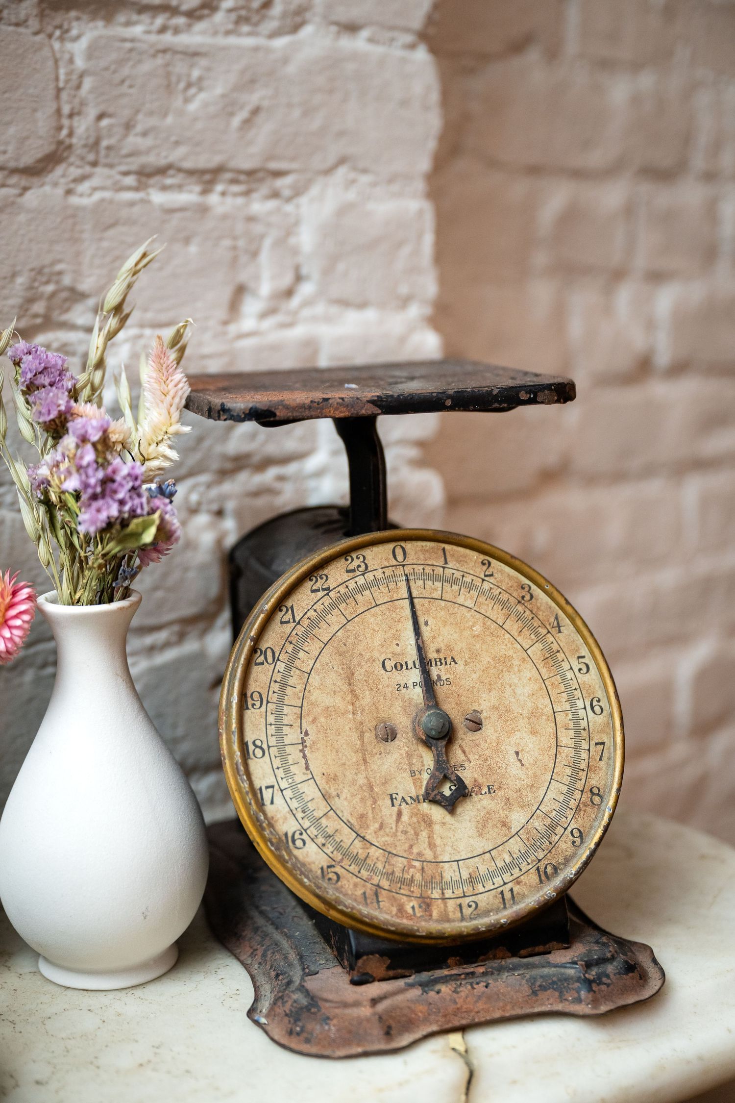Antique rustic scale with white vase of purple wildflowers against white brick wall.