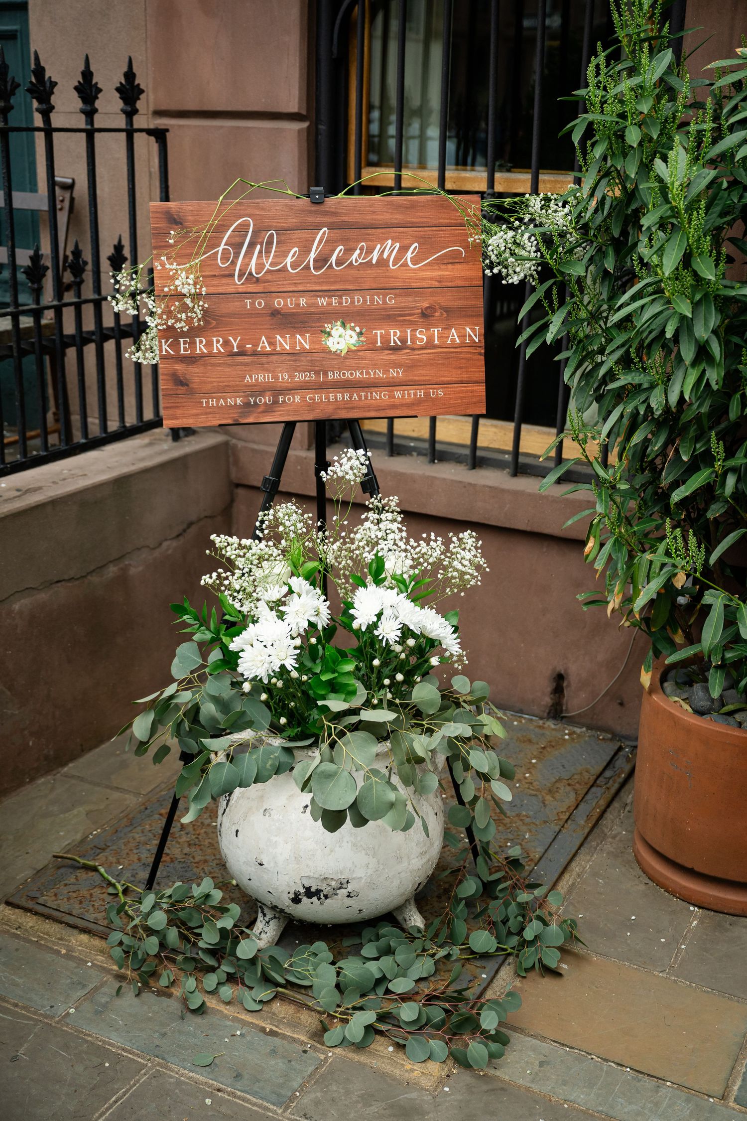 Rustic wooden welcome wedding sign with white floral arrangement in concrete planter on brownstone steps.