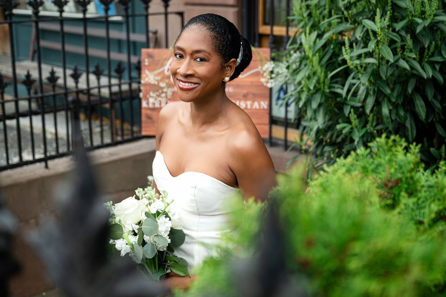 A radiant bride in a strapless white gown holds a bouquet of white flowers while smiling outdoors near greenery and iron railings.