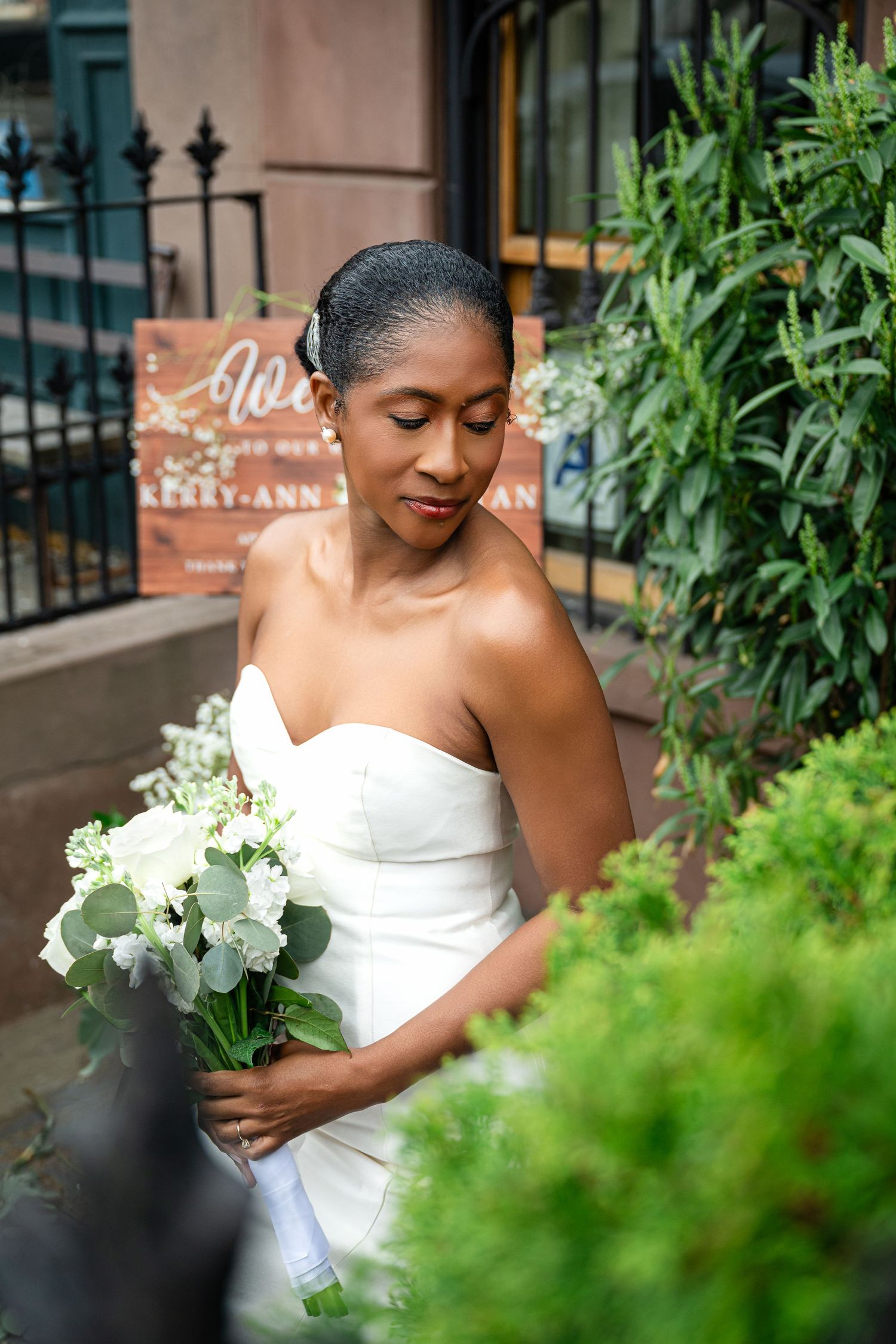 Elegant bride in strapless white gown holds a bouquet of white flowers while standing near greenery and iron railings.