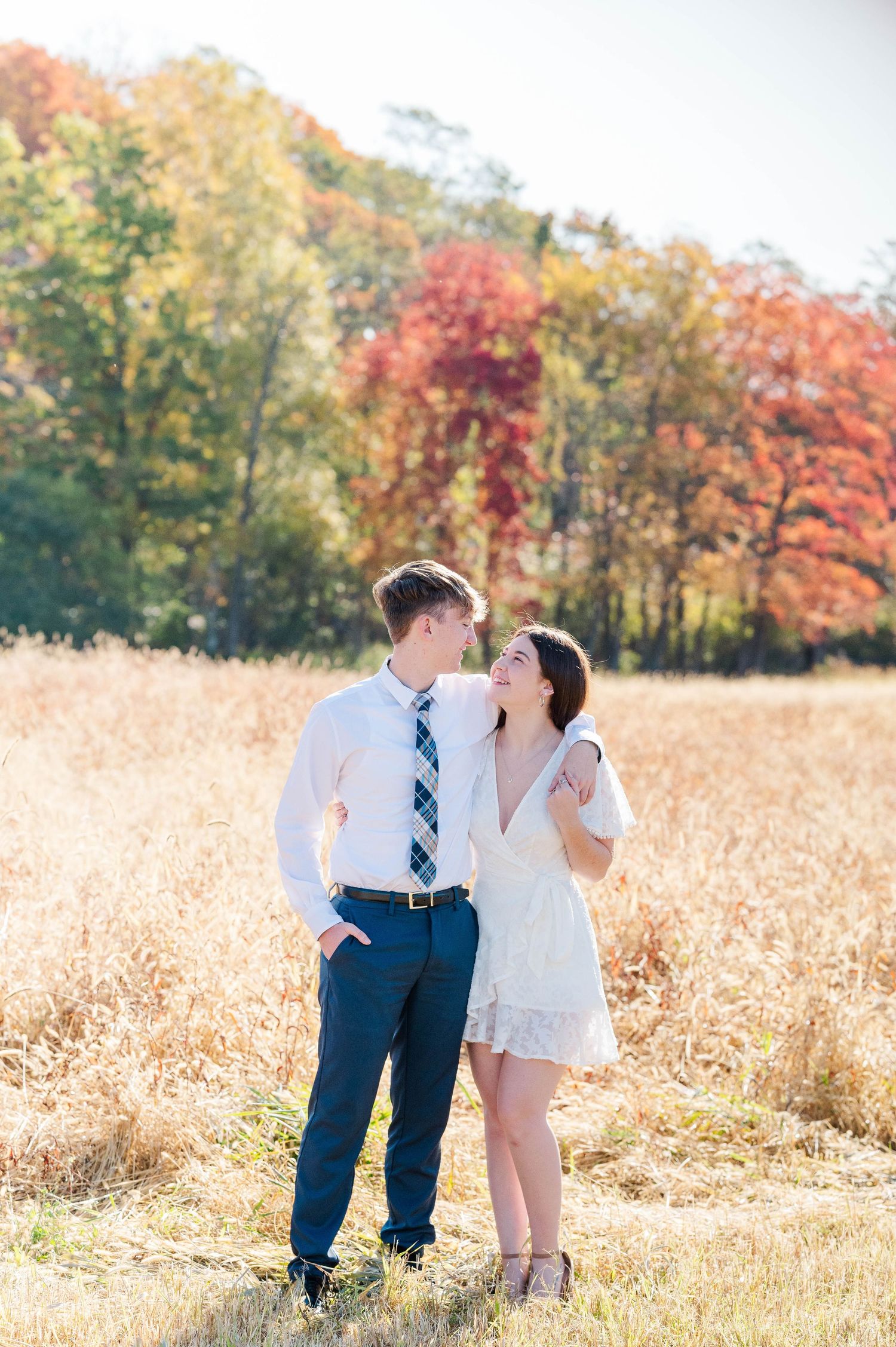 A couple shares a romantic moment in a golden wheat field with vibrant fall foliage in the background.