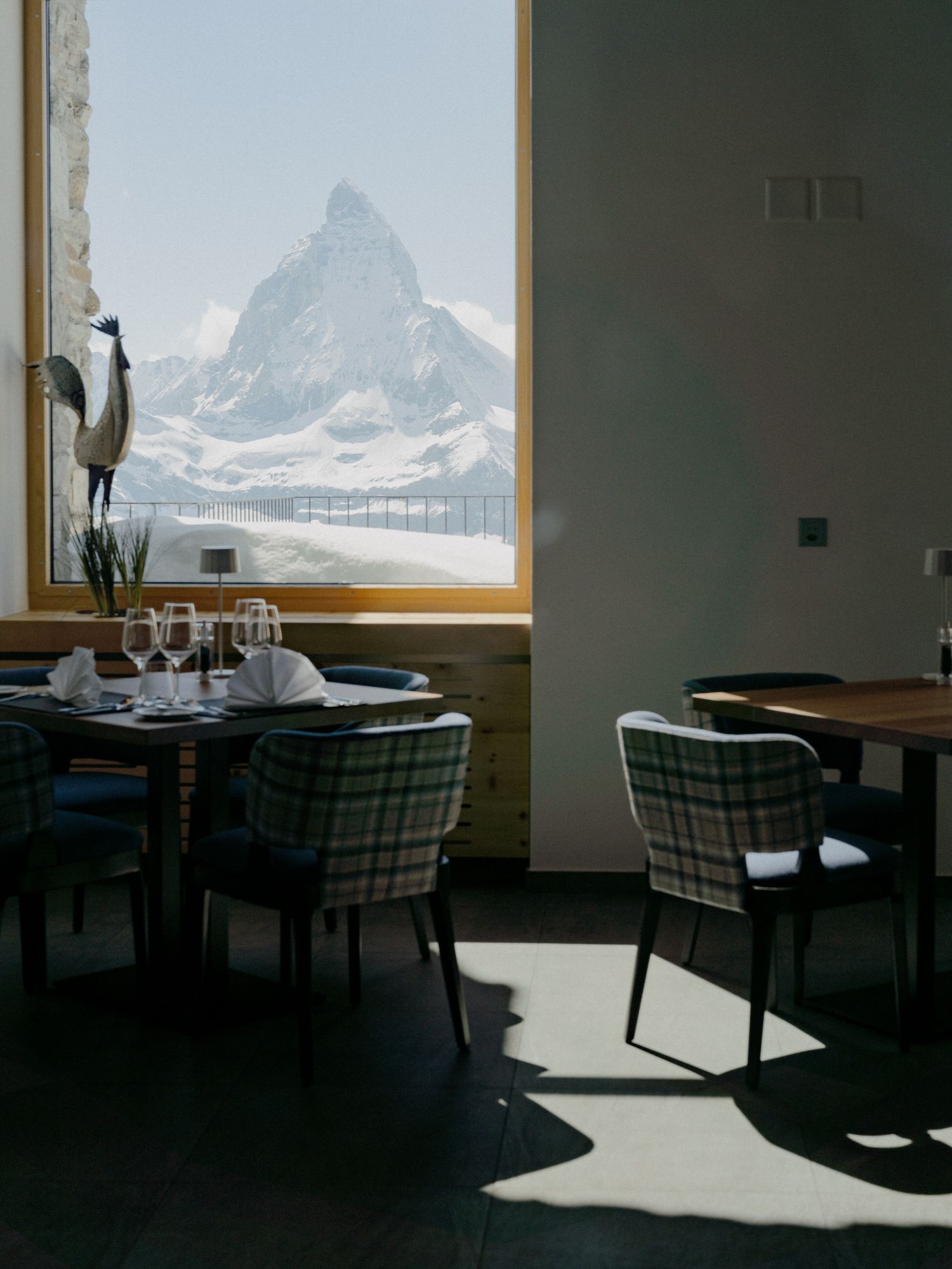 A dimly lit restaurant dining room with plaid chairs and a dramatic mountain vista visible through large windows.