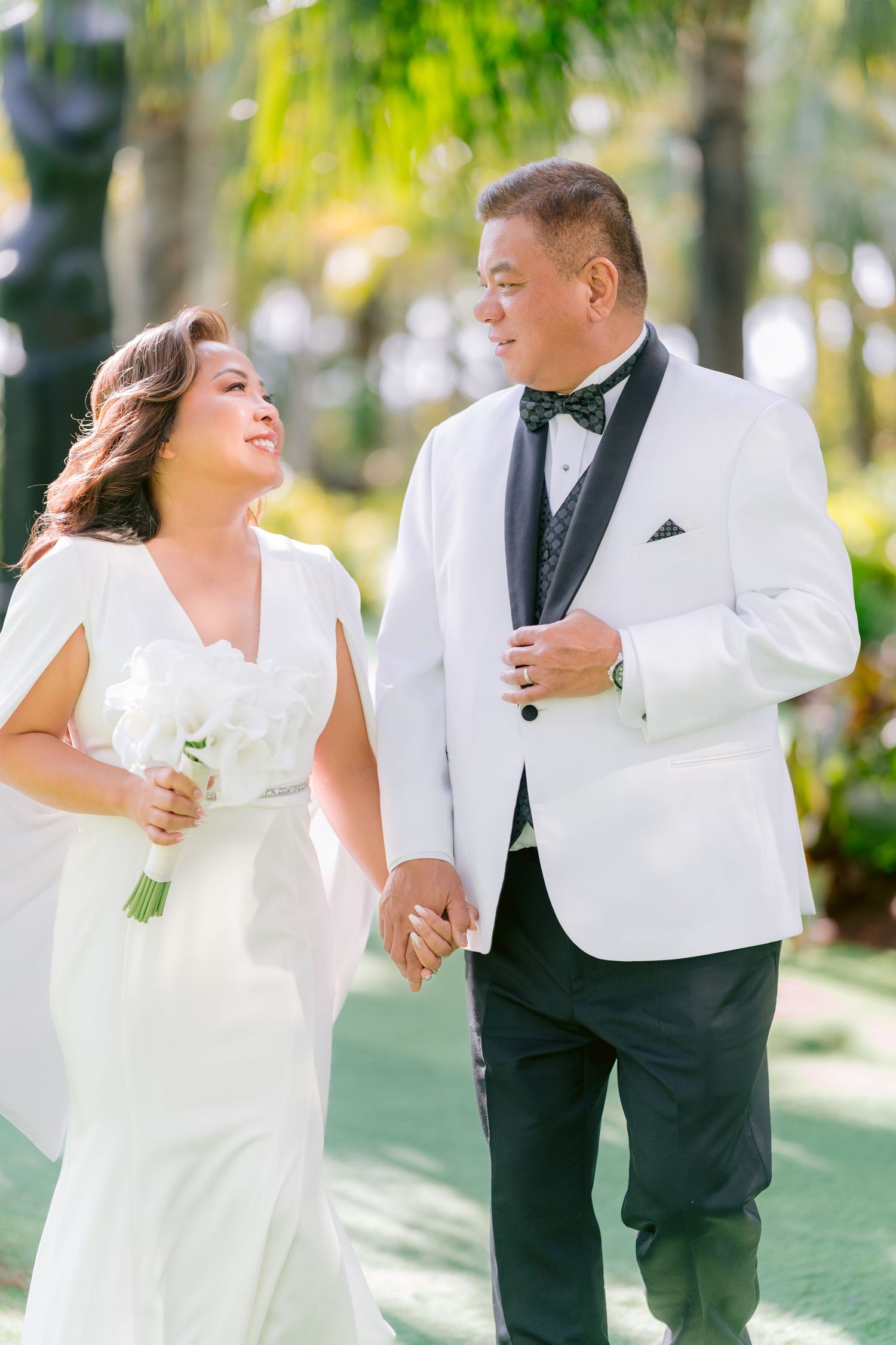 Happy newlyweds in wedding attire walking together outdoors in the Lake Nona Wave Hotel garden setting with sunlit trees in the background.