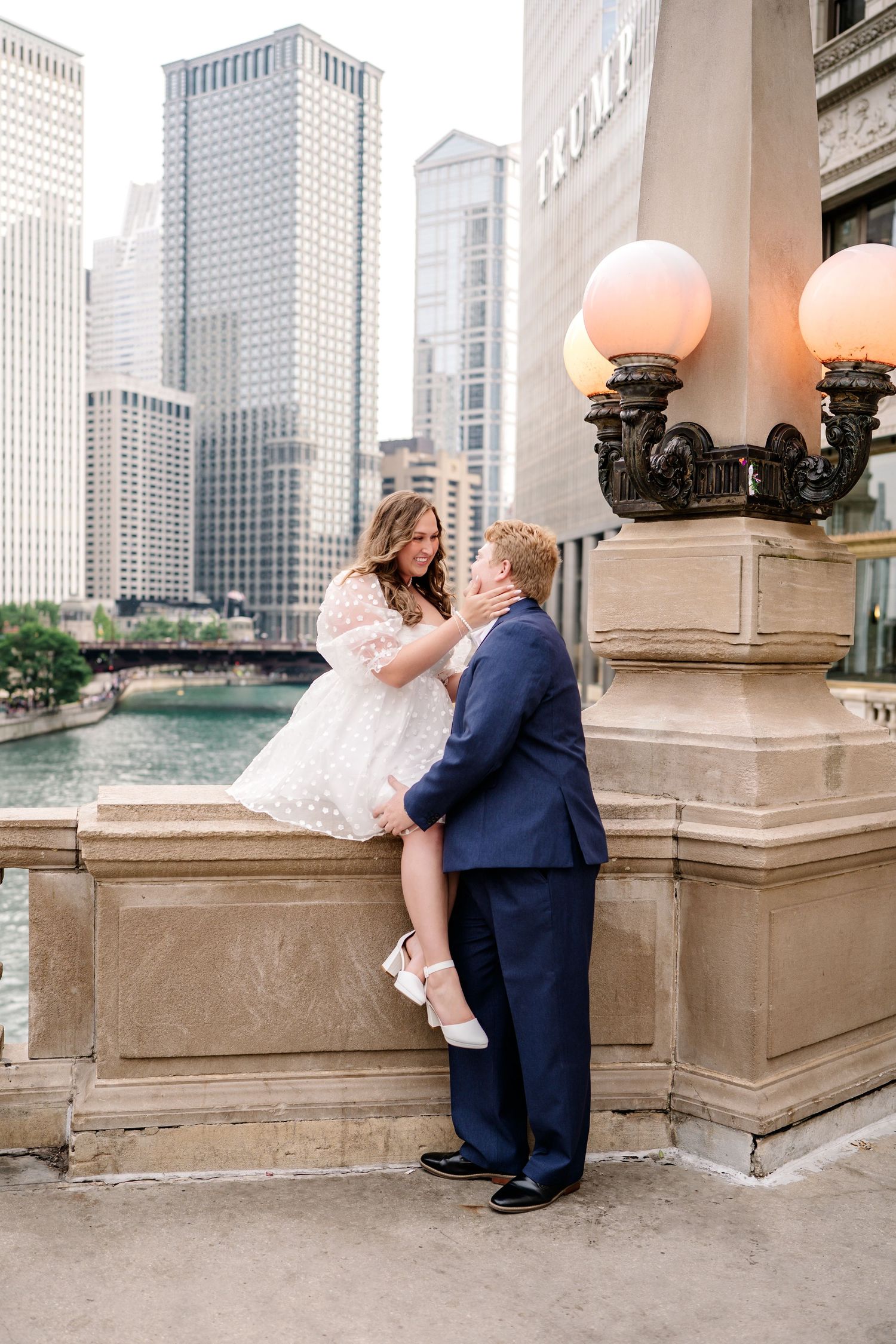 A couple embraces on stone steps with ornate lamp posts and city buildings in the background along a riverwalk.