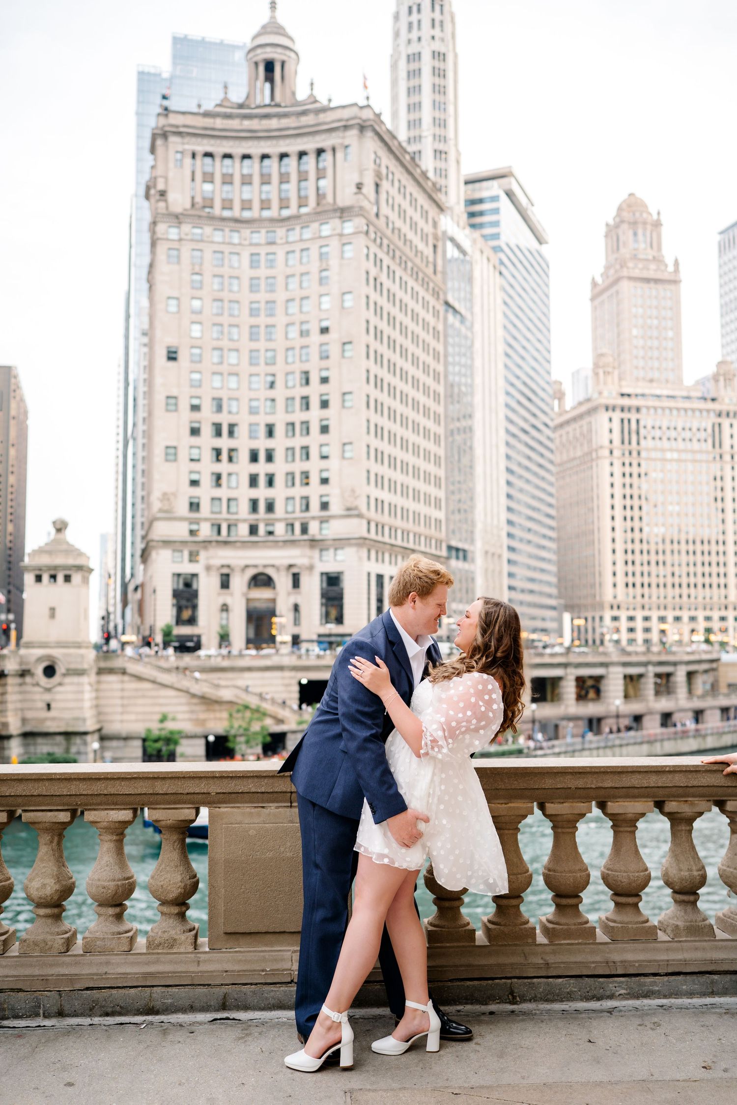 Romantic couple takes engagement photos on Chicago Riverwalk with historic Wrigley Building and downtown architecture in background.