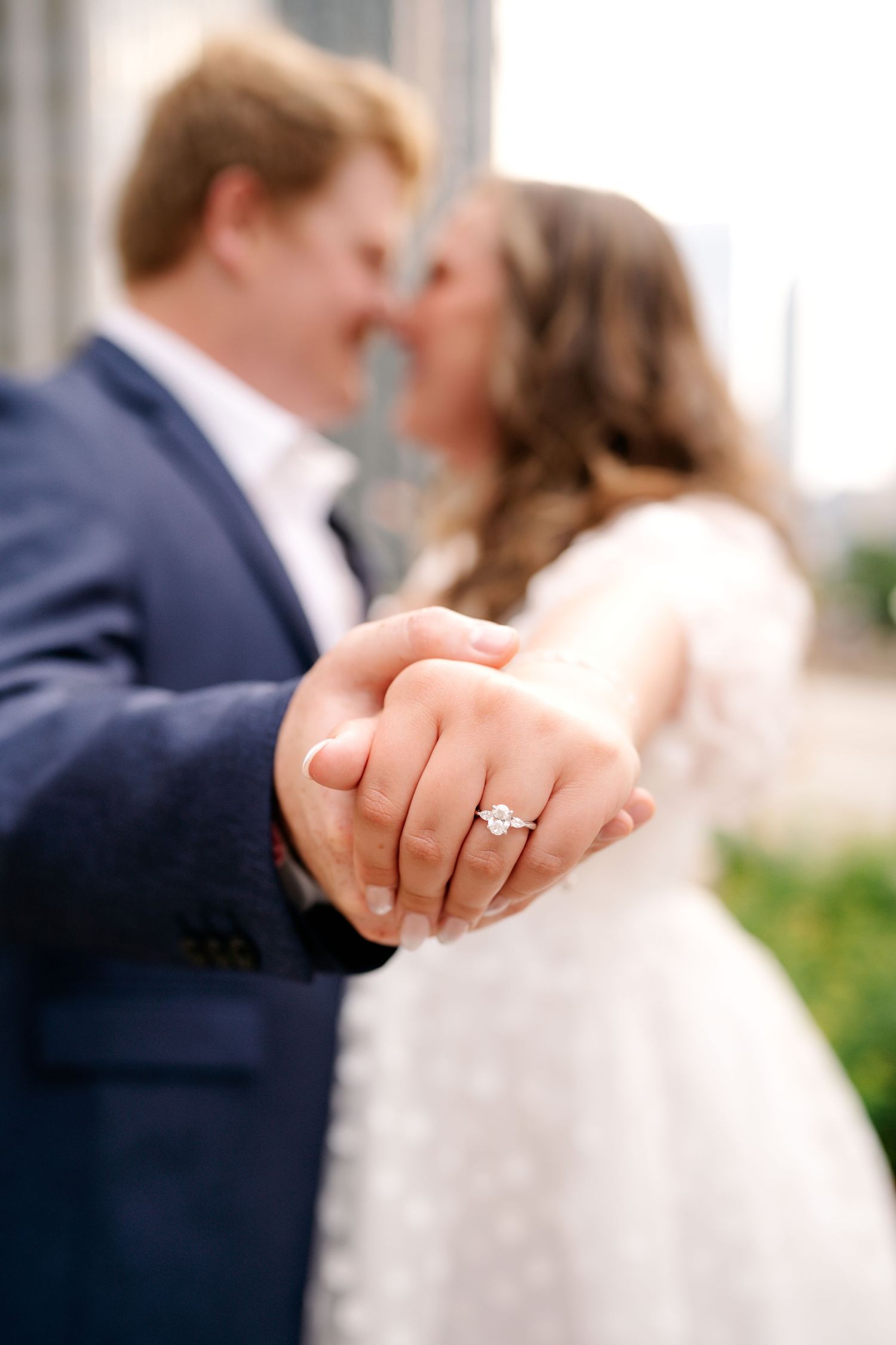 Close-up of wedding rings and joined hands with couple embracing in soft focus background.