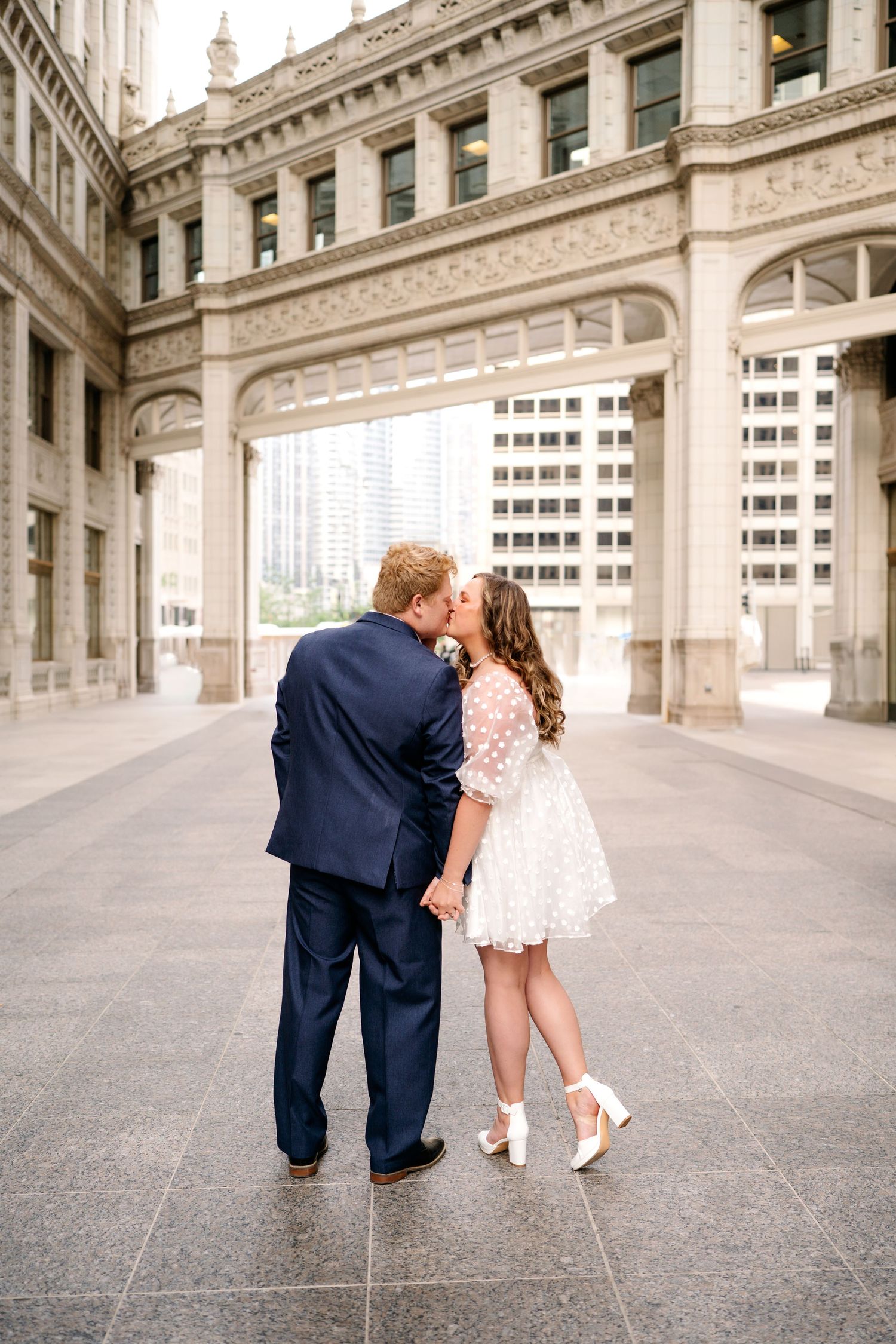 Couple shares romantic kiss under ornate stone archway in urban setting with bride wearing short white lace dress.