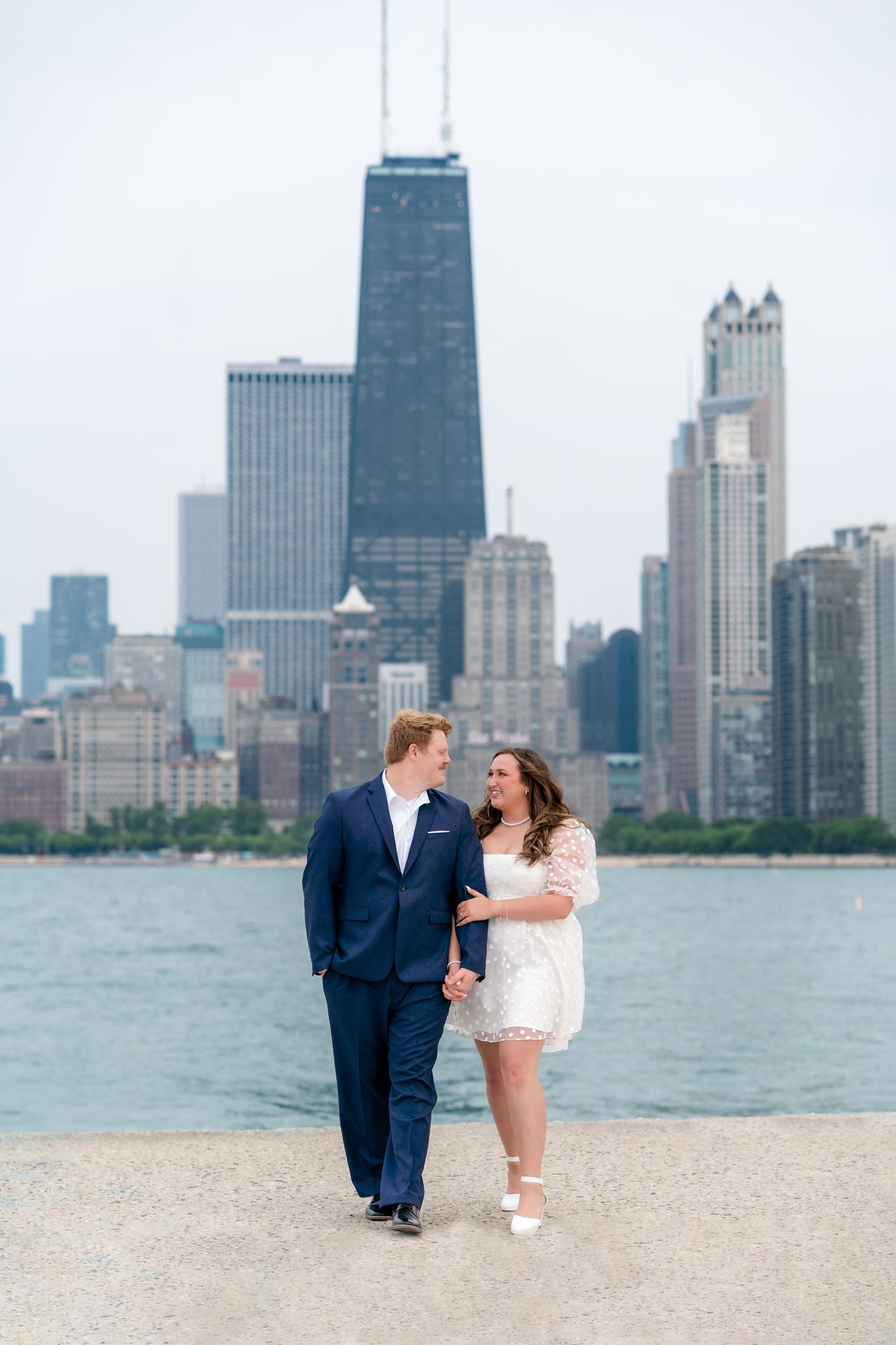 A couple walks along Lake Michigan with Chicago's iconic skyline featuring the John Hancock Center in the background.