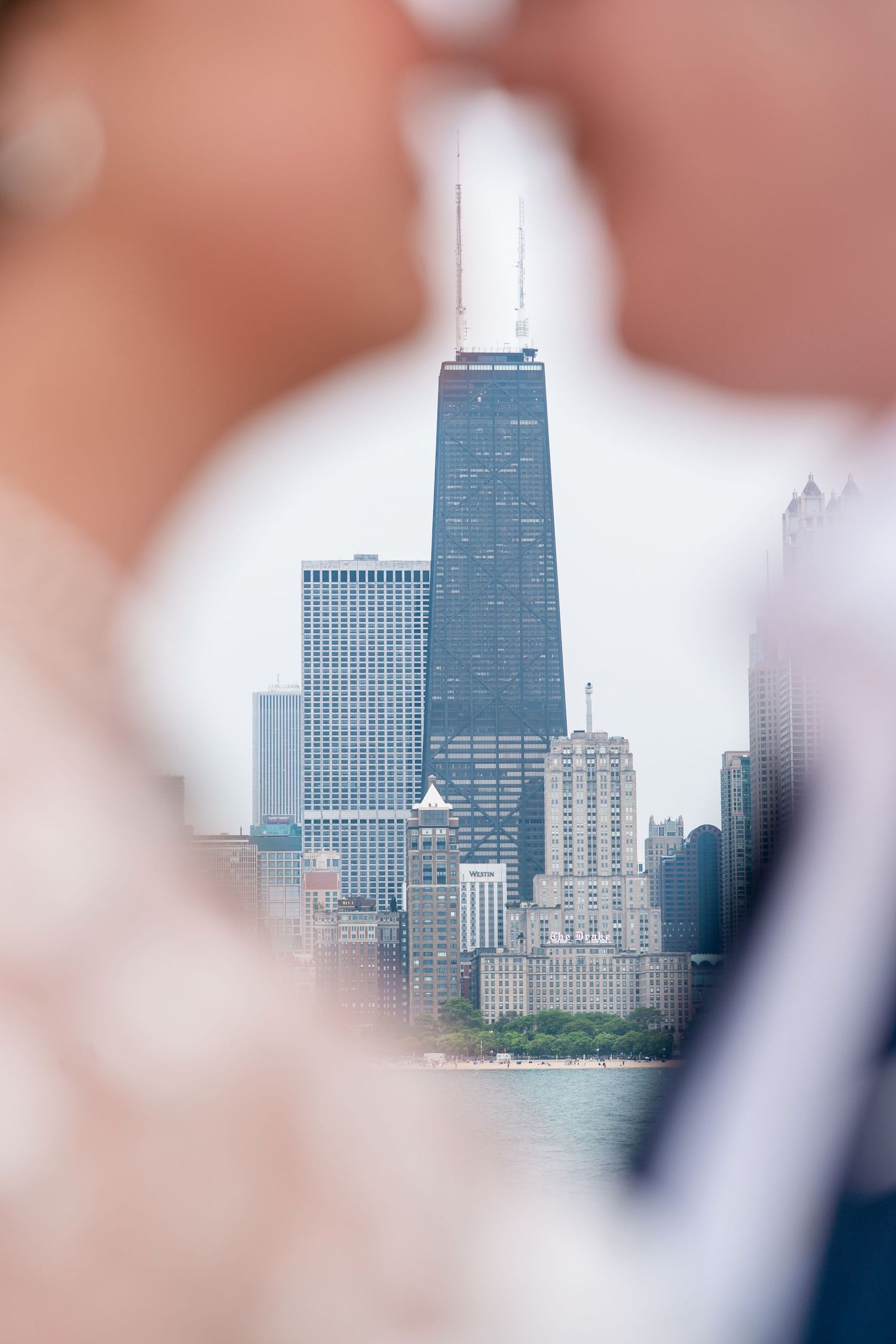 Chicago's iconic John Hancock Center and skyline viewed through a blurred foreground frame near Lake Michigan.