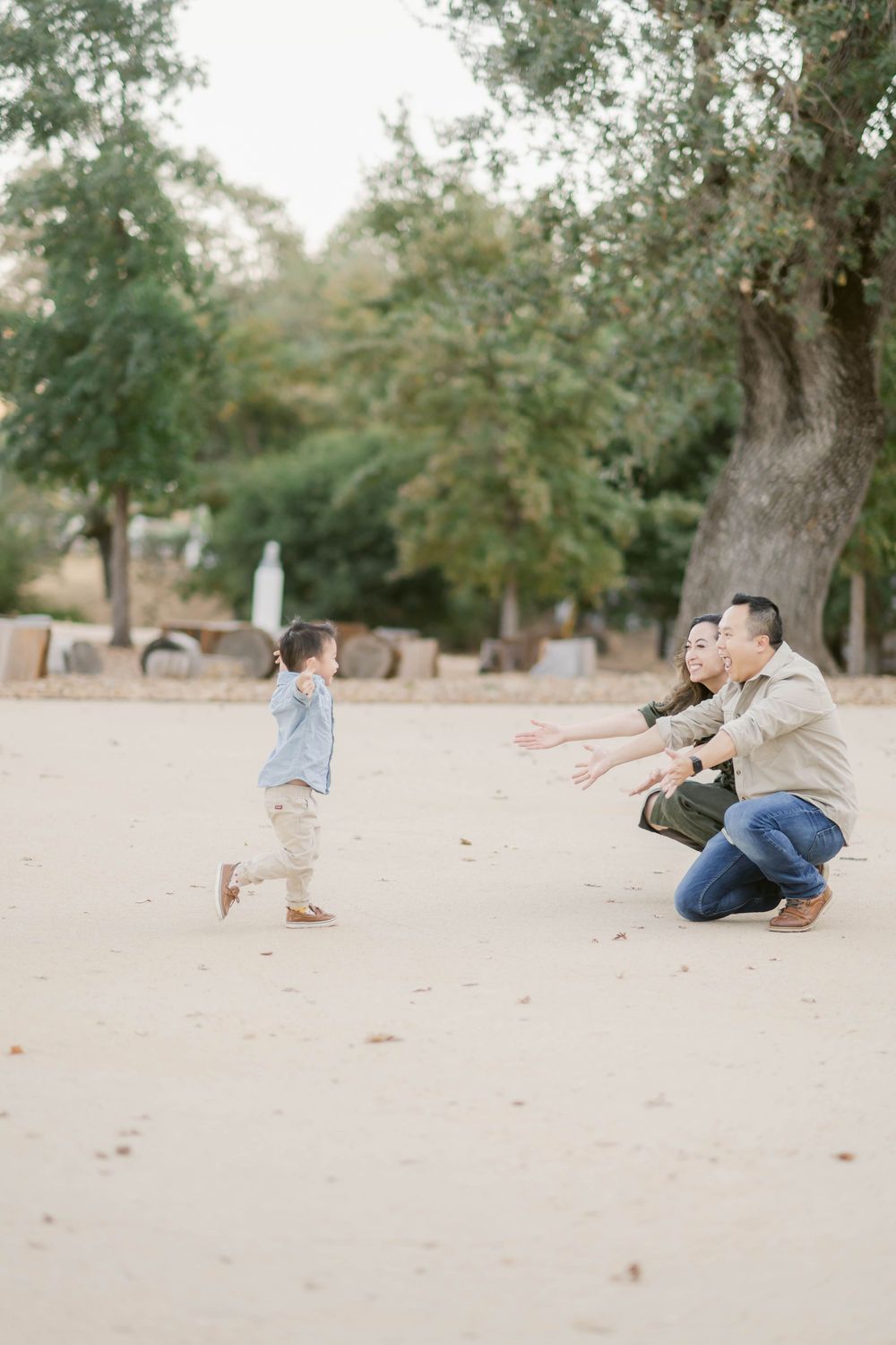 A small child runs joyfully towards two people crouching on a sandy ground surrounded by trees.