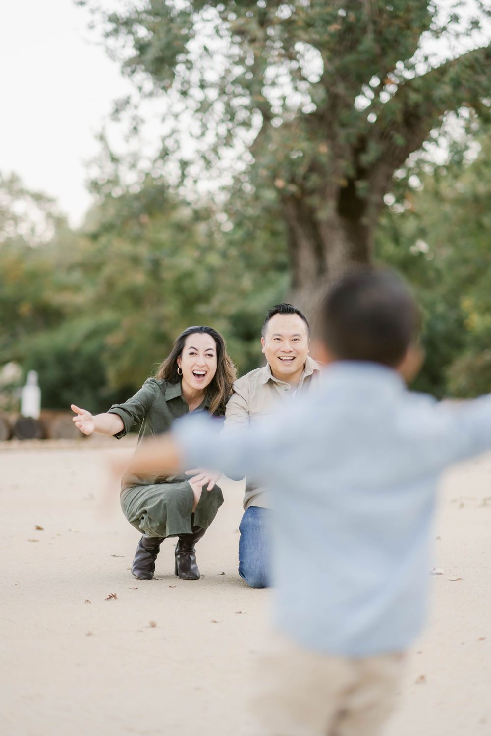 A couple crouches down and smiles joyfully at their child running towards them on a sandy path under trees.