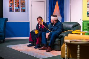 Two actors sit close together on a dark couch during a theatrical scene.