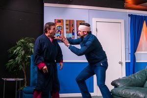 Two actors in dark clothing engage in a dramatic confrontation scene on stage.