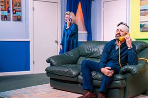 Two performers in blue outfits sit on a leather couch during a theatrical scene.