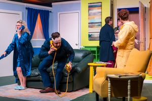 Actors perform a comedic scene in a colorful living room set with blue walls and yellow accents.