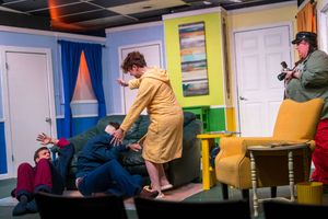 Theatrical performers engage in physical comedy on a vibrant stage set with a green couch and yellow chair.