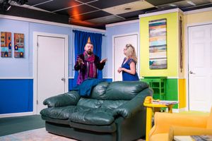 Two performers act out a dramatic scene on a dark green leather couch in a colorfully decorated room.