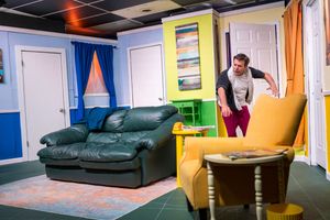 Actor leaning into a living room stage set with a green couch and yellow armchair.