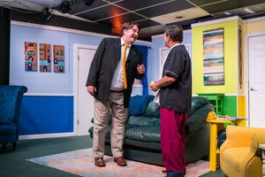 Two actors engage in dialogue during a theater performance with colorful stage design.