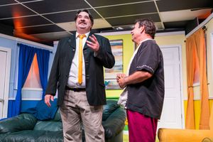 Two performers engage in dialogue during a theatrical scene on a colorful stage set.