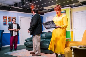 Actors in vintage clothing perform a scene in a retro-styled living room with bright blue walls.