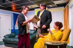 Three performers act out a scene on stage while one reviews a document in yellow attire.