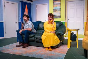 Two performers sitting apart on a green couch in a colorful stage set with bright yellow and blue walls.