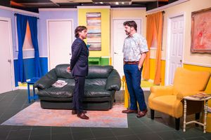 Two performers have a conversation while standing near a dark leather couch on a theatrical stage set.