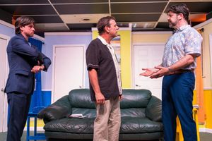 Three male actors in casual attire perform dialogue scene on stage with green couch backdrop.