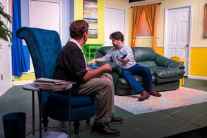 Two actors perform intimate dialogue scene on stage with leather couch and colorful walls.