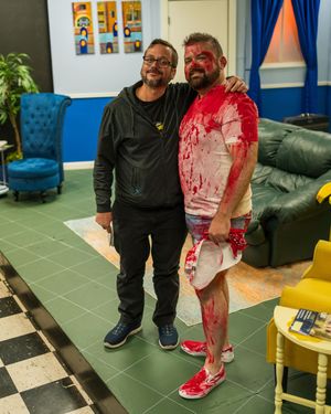 Two actors pose together on a checkerboard floor with one wearing theatrical makeup.
