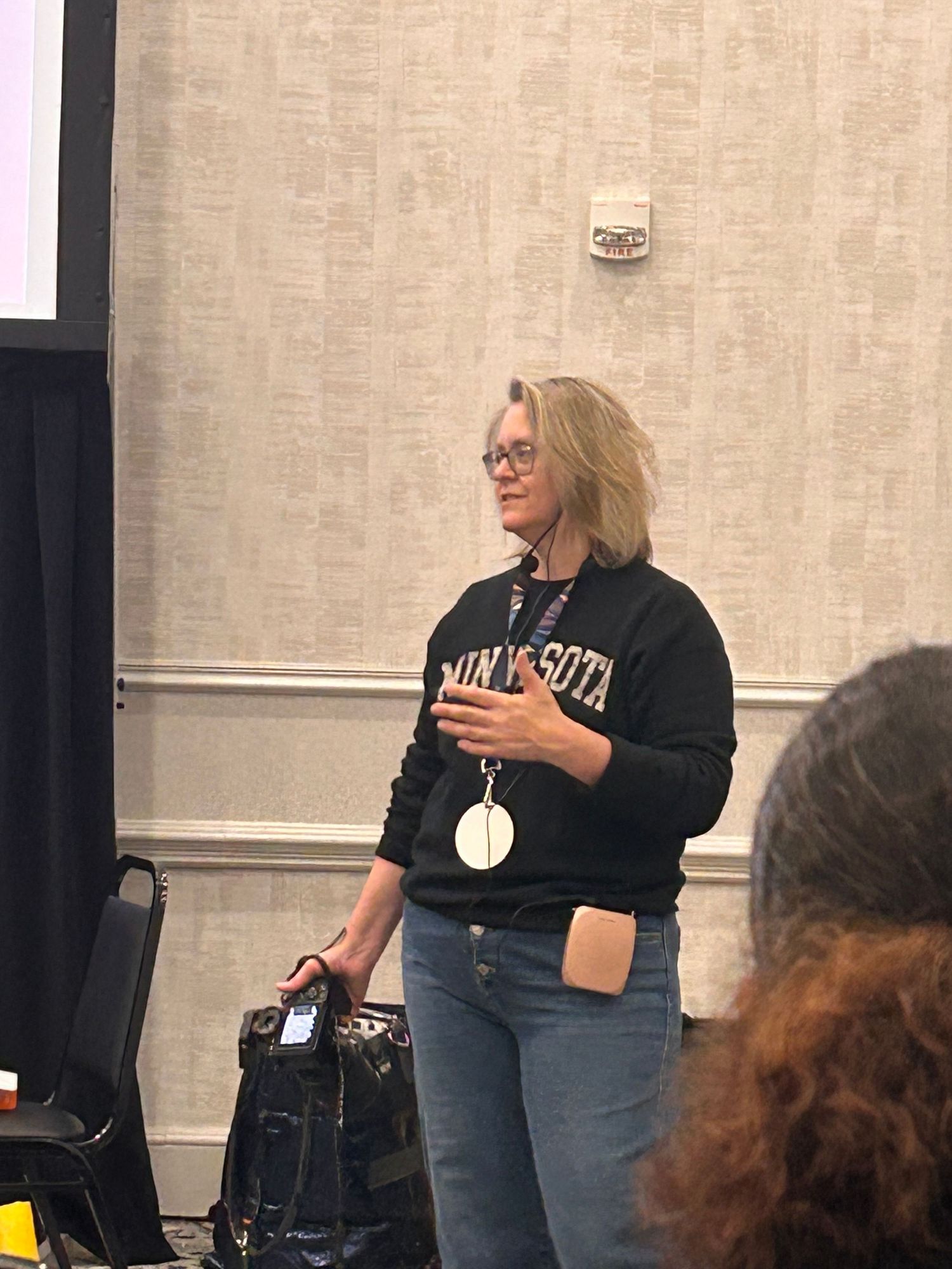 A speaker wearing glasses stands presenting to a group while dressed in a University sweatshirt and jeans.