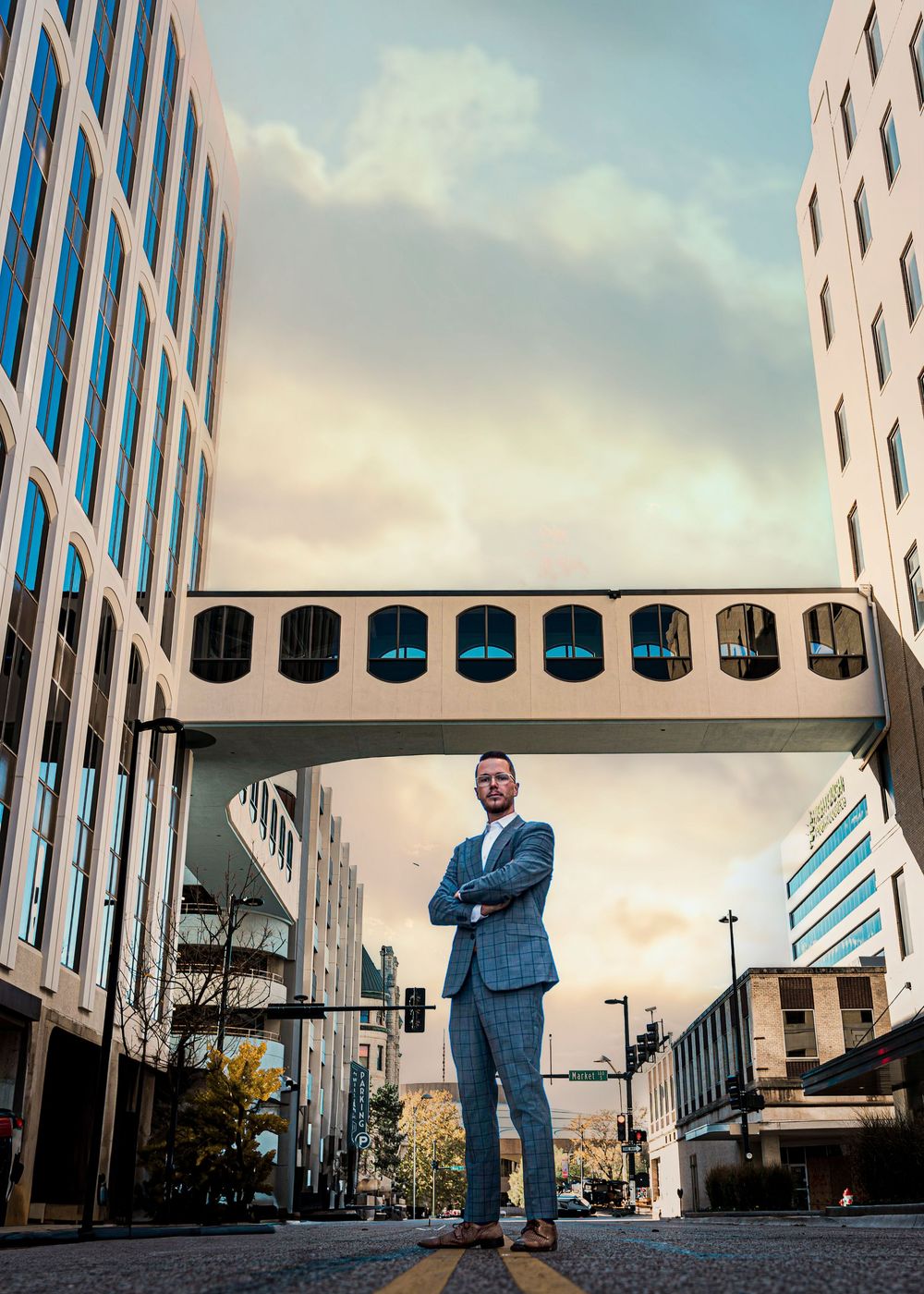 Professional in a blue suit stands between modern office buildings with a skybridge overhead in an urban setting.