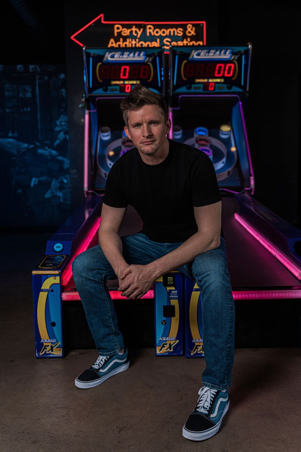 A person sits on an arcade game cabinet with neon signs and retro gaming lights in a dark arcade setting.