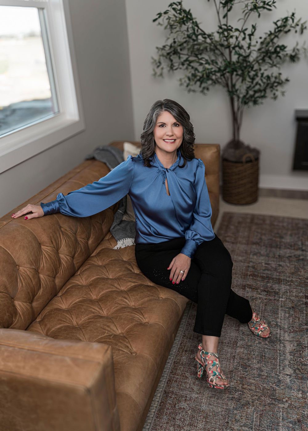 Professional woman in blue blouse sits casually on brown leather tufted couch near potted plant and window.