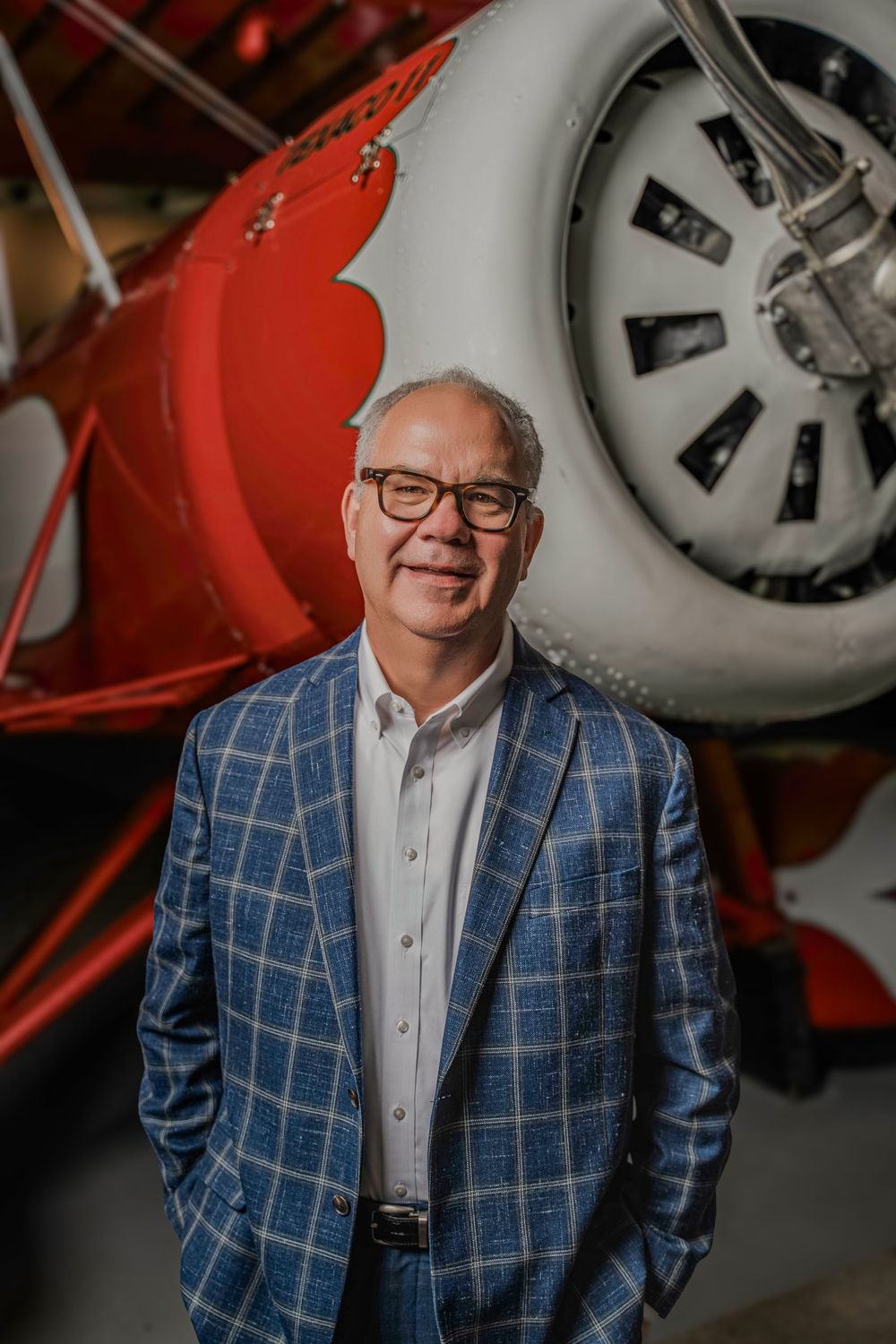 Professional wearing blue plaid blazer and glasses poses in front of vintage aircraft in aviation museum setting.