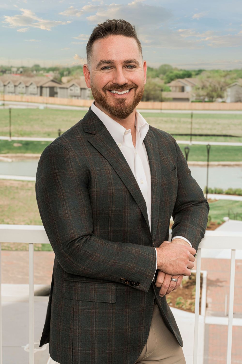 Professional portrait of man in plaid blazer and white shirt posing outdoors with architectural details behind.