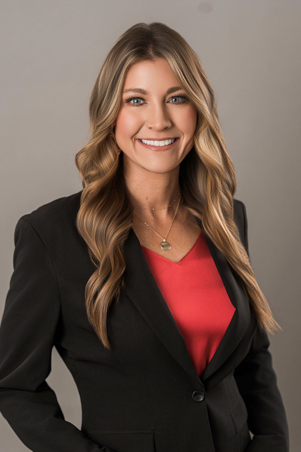 Professional business portrait with long wavy hair, black blazer and red top posing against neutral background.
