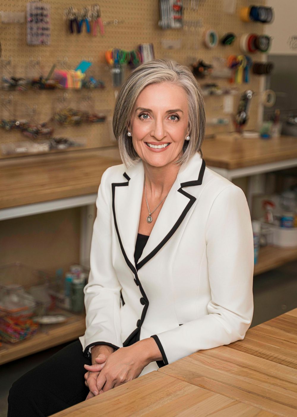 Professional in a crisp white blazer with black trim sits at wooden desk in workspace with tools and supplies on wall shelves.