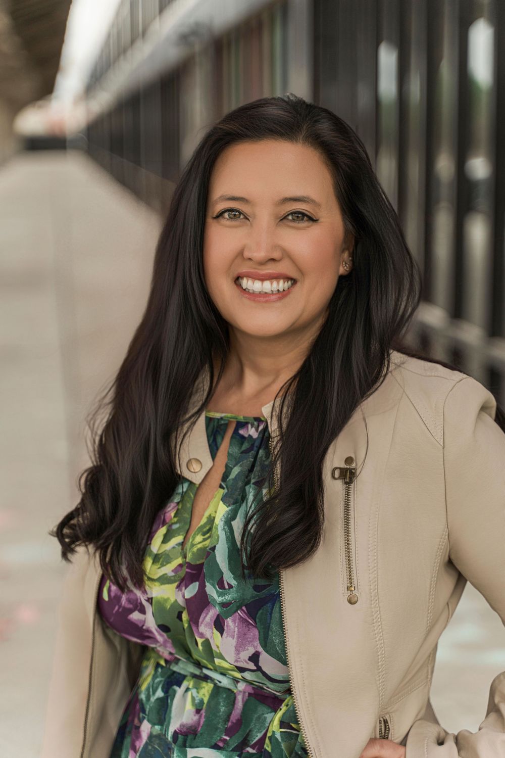 Professional headshot of a woman in a floral dress and beige blazer smiling warmly against a blurred background.