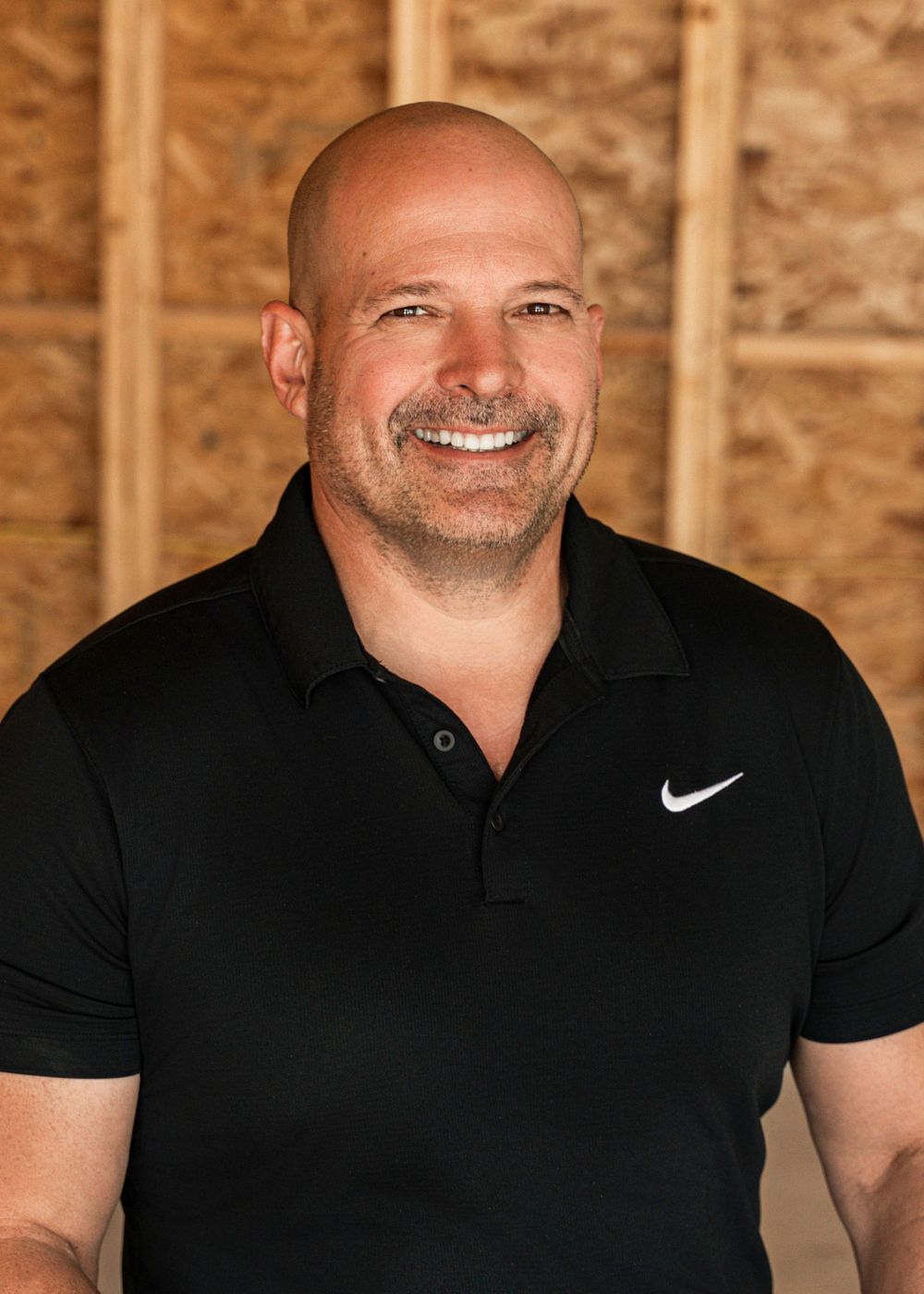 A bald person wearing a black Nike polo shirt smiles warmly at the camera against a wood panel background.