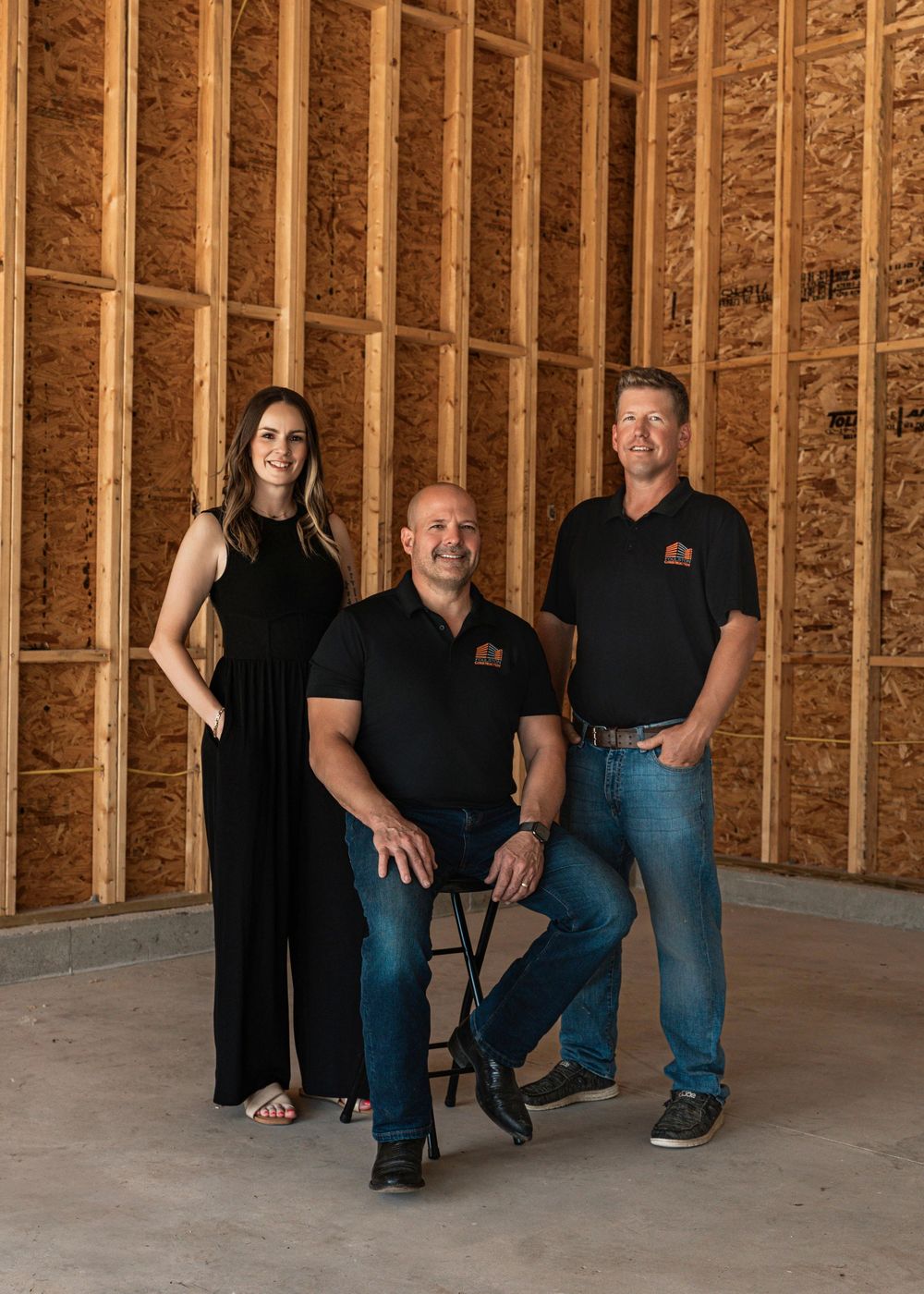 Three employees wearing black polo shirts and jeans pose together in an unfinished room with exposed wooden beams and studs.