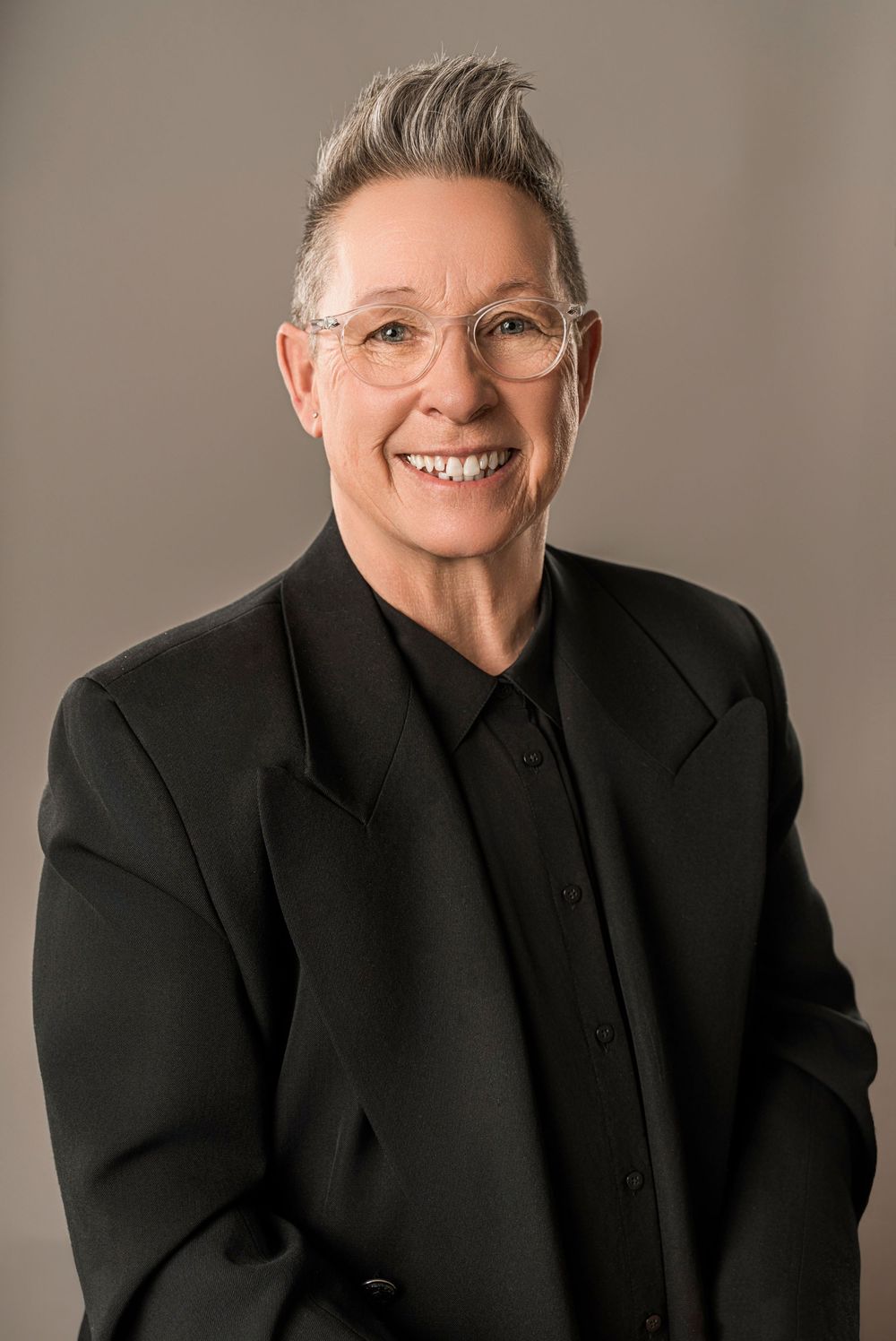 Professional portrait of a person in a black blazer and button-up shirt wearing round glasses with short spiked gray hair.