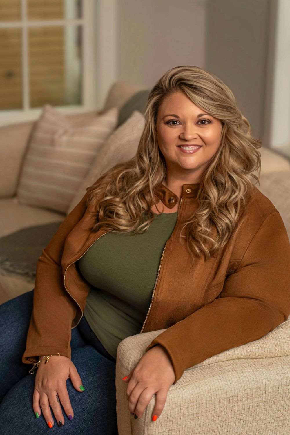 A woman with wavy blonde hair relaxes on a beige couch wearing a brown jacket and olive green top.