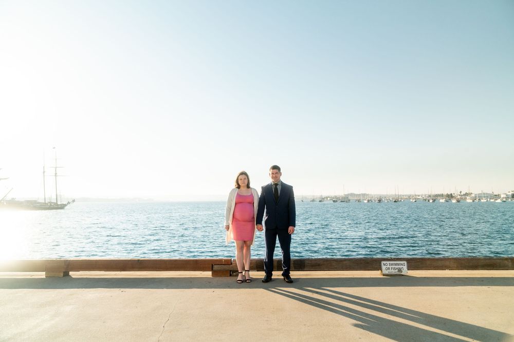 A couple stands together on a pier overlooking a serene harbor with boats and calm water on a sunny day.