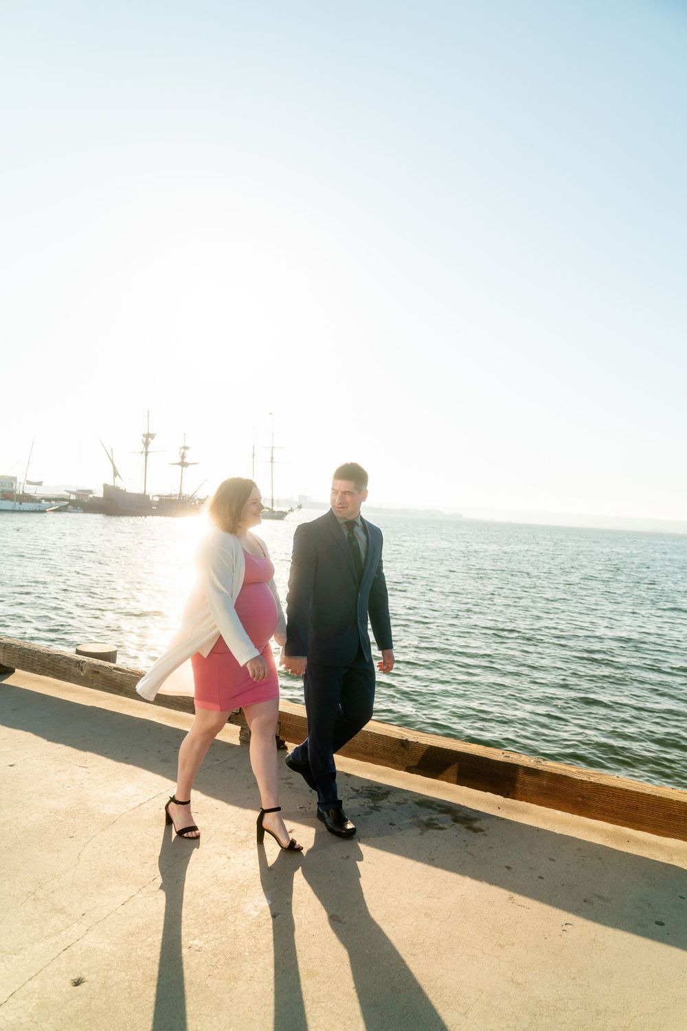 A couple walking together on a waterfront pier during sunset with a ship silhouetted in the background.
