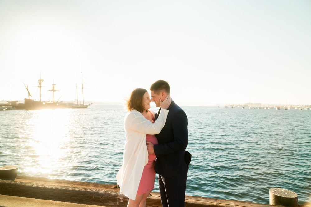 A couple shares an intimate moment at sunset along the waterfront with calm blue waters and distant boats in view.